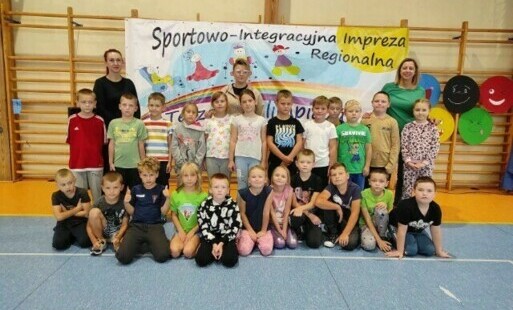 Grupa dzieci pozuje do zdjęcia z opiekunkami, w tle drabinki i plakat Sportowo-Integracyjna Impreza Regionalna